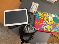 Samsung tablet, Canon camera in carrying case, battery charger in packaging, two colorful pouches on table