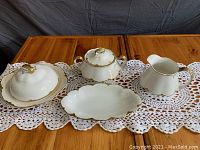 Overview of four white porcelain dishware pieces with 18k gold rims on crocheted lace mat on wood table.