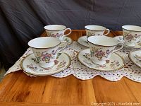 Front view of six tea cups and saucers arranged on a crocheted cloth on wooden table