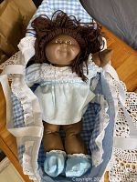 Front view of the doll lying in the blue and white checked bassinet with lace edges, showing the doll's face and outfit.
