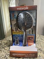 Front view of new Waterpik PowerPulse UltraThin+ hand shower head in packaging showing chrome shower head, wall mount bracket, and branding.