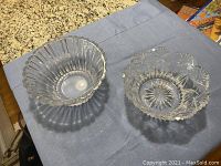 Two clear glass decorative bowls displayed on a blue cloth; one bowl with vertical ribs and scalloped rim, the other with floral cut-glass patterns and starburst base design.
