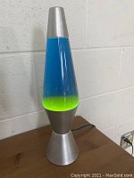 Photo of a blue and green lava lamp about 15 inches tall with a silver metallic base and cap on a wood surface against a white wall.
