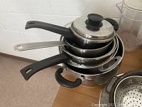 Four metal pots and pans stacked on a wooden surface, two with lids and black handles.