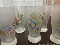 Photo showing six vintage frosted glass water glasses arranged on a table, each featuring unique hand-painted floral patterns including blue, pink, and red flowers with green leaves. The glasses have frosted rims and bases.