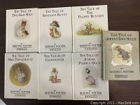 Photo showing front covers of seven Beatrix Potter books arranged in two rows on a wooden surface: The Tale of Two Bad Mice, The Tale of Benjamin Bunny, The Tale of The Flopsy Bunnies, The Tale of Mrs. Tittlemouse, The Tailor of Gloucester, The Tale of Jemima Puddle-Duck, The Tale of Johnny Town-Mouse.