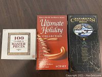 Photo showing front covers of the 3 CD collections laid out side by side on a wood floor. Includes '100 Classical Masterpieces', 'Ultimate Holiday Collection' 6 CD box, and 'Ultimate Christmas Volume 2' single CD.