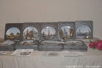 View of nine Limoges porcelain collector plates featuring Paris France scenes by Louis Dali, each plate includes a decorative frame and is displayed standing on table.
