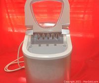 Open electric countertop ice maker showing interior with ice molds and power cord to the side against red backdrop.