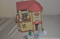 Front angled view of Calico Critters Country Cottage House dollhouse showing the red roof, beige walls, front door, and balcony with several Paw Patrol figures displayed. Additional figures and packaging placed in front of the dollhouse.