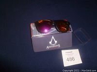Assassins Creed sunglasses resting on original branded metal case with logo visible.