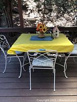 Outdoor set showing the table covered with a yellow tablecloth and four white wrought iron chairs with cushions surrounding it on a wooden deck.