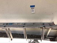 Full side view of aluminum metal ladder against a light-colored wall, showing rungs, side rails, and stability feet.