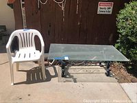 Photo of white plastic outdoor chair and rectangular clear glass table with wrought iron base placed outside on concrete surface.