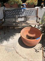 Wood and metal outdoor bench with decorative lattice backrest, wooden slats for seat and back, alongside a large terracotta clay pot on concrete surface with planters in the background.