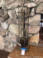 Full view of brass fireplace tool set with six tools on brass stand in front of stone fireplace.