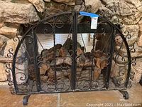 Front view showing full fireplace screen with arched top and scrollwork, mesh behind, standing in front of stone fireplace with wood inside.