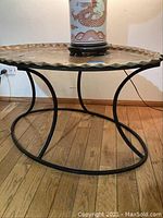 Full view of brass table showing scalloped edge and black curved metal base on wood floor.