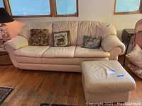 Full view of cream leather sofa with three throw pillows and matching ottoman placed on hardwood floor in a living room setting.