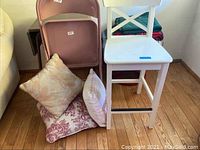 Photo showing pink folding chair, white counter chair, decorative pillows on floor, and some folded blankets behind.