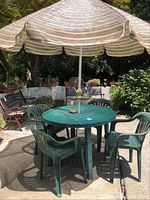 Outdoor setup showing round green plastic table with one object on top, four matching matching plastic chairs around it, and a large striped umbrella overhead providing shade.