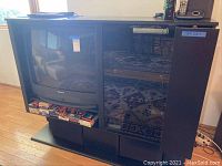 Front view of black woodgrain entertainment center with built-in speaker on the right side and space for CRT TV on the left. VHS tapes stored on bottom shelf.