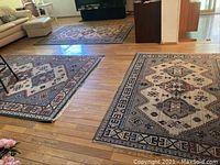 Photo showing two large woven area rugs with geometric and ethnic motifs on hardwood floor in living room space, arranged near couches and furniture.