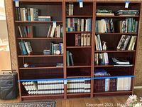 Three large wooden bookshelves filled with assorted hardcover and paperback books; bottom shelves contain encyclopedias not included in lot.
