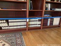 Lower two shelves of a wooden bookshelf with uniform volumes of encyclopedias with light covers and other books arranged horizontally and vertically on hardwood flooring next to a patterned rug.