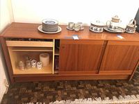 Sideboard with ceramic dishware including bowl, mugs, assorted plates, candles, candle holders, and napkin rings arranged on top and inside open compartment