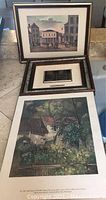 Three framed wall art prints showing various landscapes and street scenes, some with people, arranged on a surface.