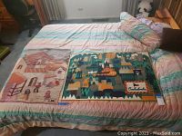 Two framed textile art pieces laid on a bed; one is a rustic house tapestry, the other a Czechoslovakia-themed map