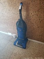 Photo of blue Hoover upright vacuum cleaner against a wall, cord wrapped around handle, showing scratches and wear.