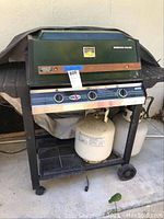 Full frontal view of green Barbecues Galore propane BBQ grill with control knobs, propane tank below, and cover partially draped over the top.