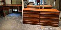 Photo showing a wooden desk with drawers on the left side and a wood dresser with multiple drawers placed in front of a mirror.