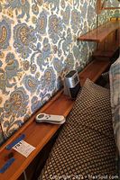 Close-up showing wooden bed frame side shelf with remotes and small device on top, set against floral wallpaper.