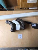 Full view of white downspout pipe and three downspout elbows (two brown, one white) on table with auction lot tag 85.