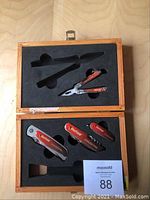 Wooden case with foam insert holding red-handled knives and multi-tool pliers.