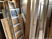 Photo showing old metal ladder leaning against plywood, white mouldings and trim pieces and various lumber pieces standing upright.