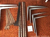 Metal shelf brackets in different sizes and colors arranged on an orange carpet. Metal rails placed in between the brackets.