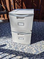 Front view of light gray plastic two drawer file cabinet placed on outdoor patterned rug. Shows top and front with handles, label slots, and locking key cylinder.