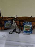 Two new 14-inch black metal hanging baskets with leaf design and coconut fiber liners attached, displayed on a white surface.