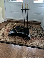 Full view of the black metal log holder with curved gold-toned handles and the fireplace tools assembled on the base showing brush, shovel, and poker.