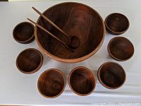 Full view of large teak salad bowl with the seven smaller bowls arranged around it, including wooden fork and spoon inside the large bowl.