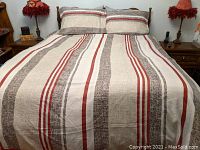 Queen size bed with striped comforter and two matching pillowcases featuring red, beige, and brown colors, displayed on wooden bed frame with wooden side tables and lamps.