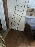Bronze coloured metal towel rack leaning against a wall on wooden floor showing overall frame and support structure.