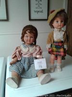 Two vintage dolls, one seated French bisque doll 22 inches tall wearing pink blouse and blue striped skirt; standing doll 17 inches tall wearing mustard jacket, plaid skirt, and a beret hat.