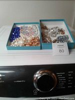 Two trays of assorted costume jewelry placed on top of a washing machine. Trays contain pearl necklaces, blue bead necklace, and metallic brooches and earrings.