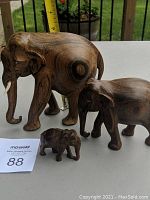 Three wooden elephant figurines arranged by size on a table with a measuring tape behind them to show scale. Largest elephant has tusks visible.