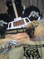 Various Hamilton Tiger-Cats fan apparel and accessories displayed together, showing t-shirts, foam finger, pom-poms, and signage.